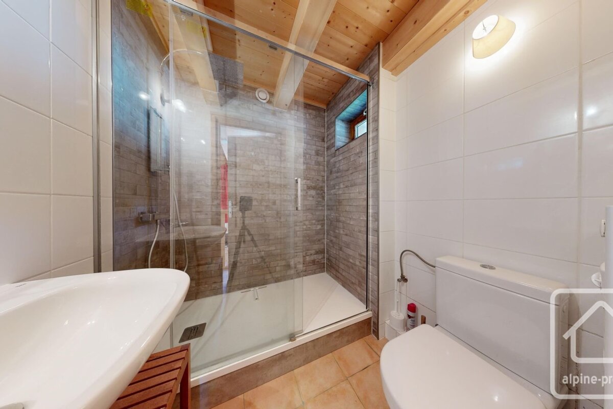 A modern bathroom features a glass shower with brick-look tiles, a white sink, and a toilet. It has white tiled walls, a wooden beamed ceiling, and brown floor tiles.