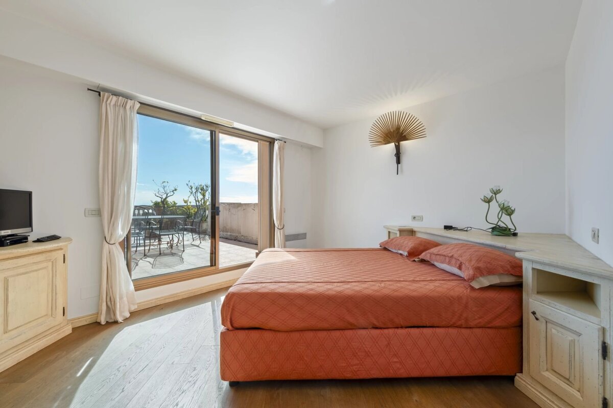 Bright bedroom with an orange bed, white walls, and a wooden floor. Sliding glass doors open to a balcony with outdoor seating and a view.