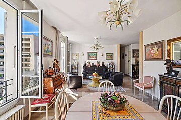 A traditional living/dining room with a black leather sofa, ornate wooden furniture, multiple paintings, chandeliers, and an open balcony door.