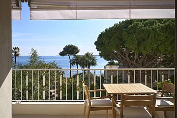 A sunny balcony with a wooden dining set and awning offers a stunning view of the blue sea, lush trees, and palm trees under a clear sky.