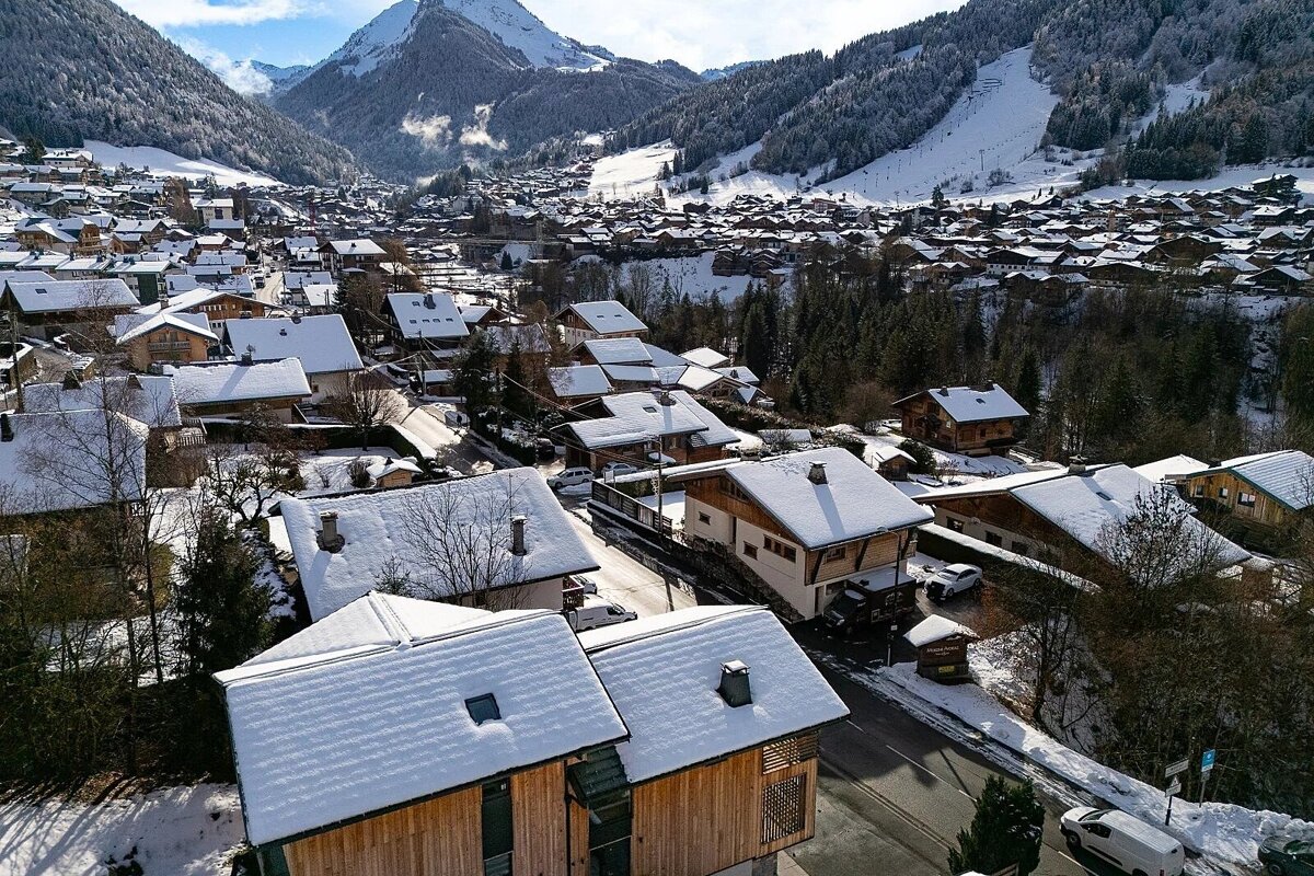 Aerial view of a charming snow-covered village in a mountain valley, with cozy chalets and towering peaks under a clear sky.