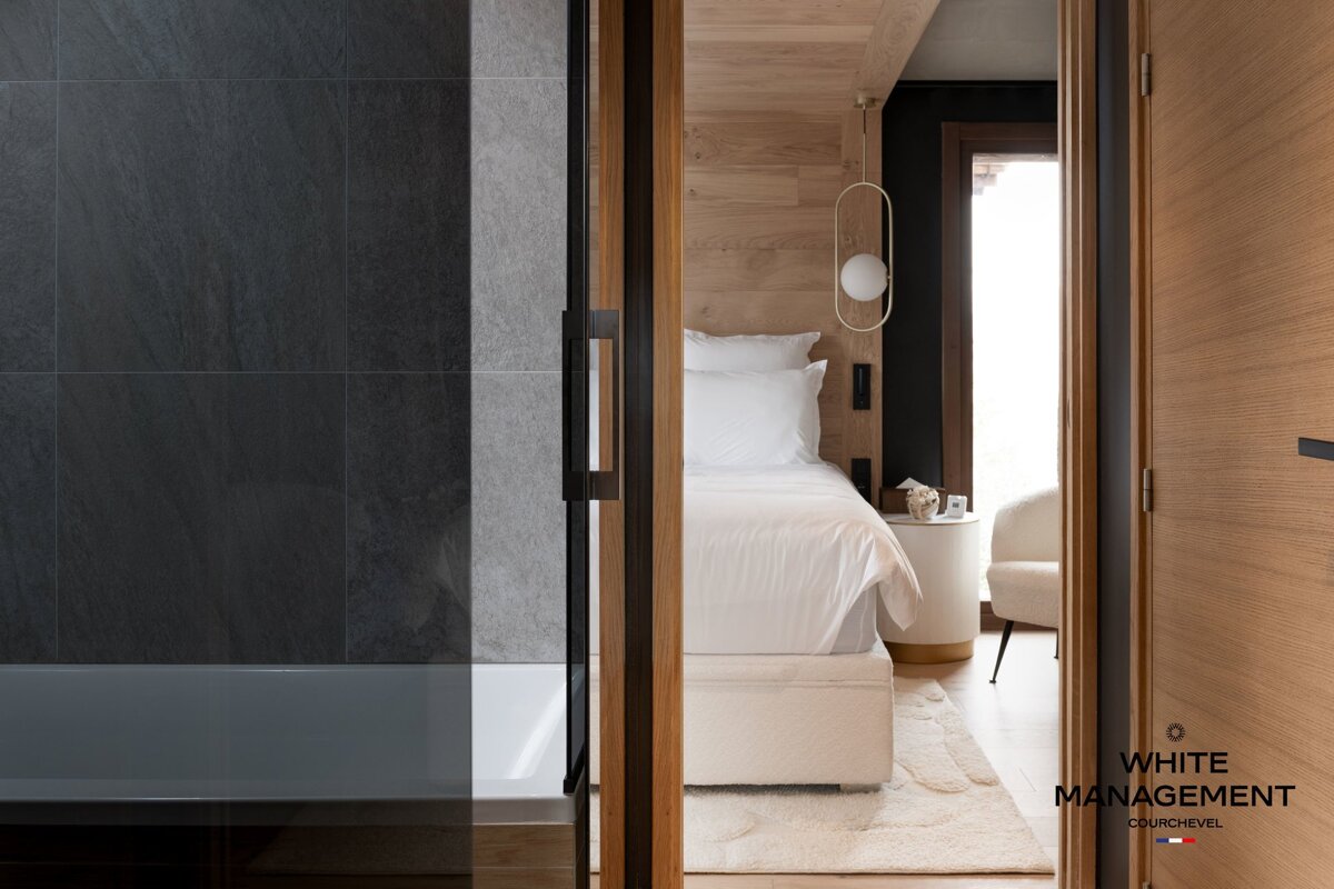 View from a sleek dark-tiled bathroom into a modern bedroom with a white bed, wooden accents, and contemporary lighting, divided by a glass panel.