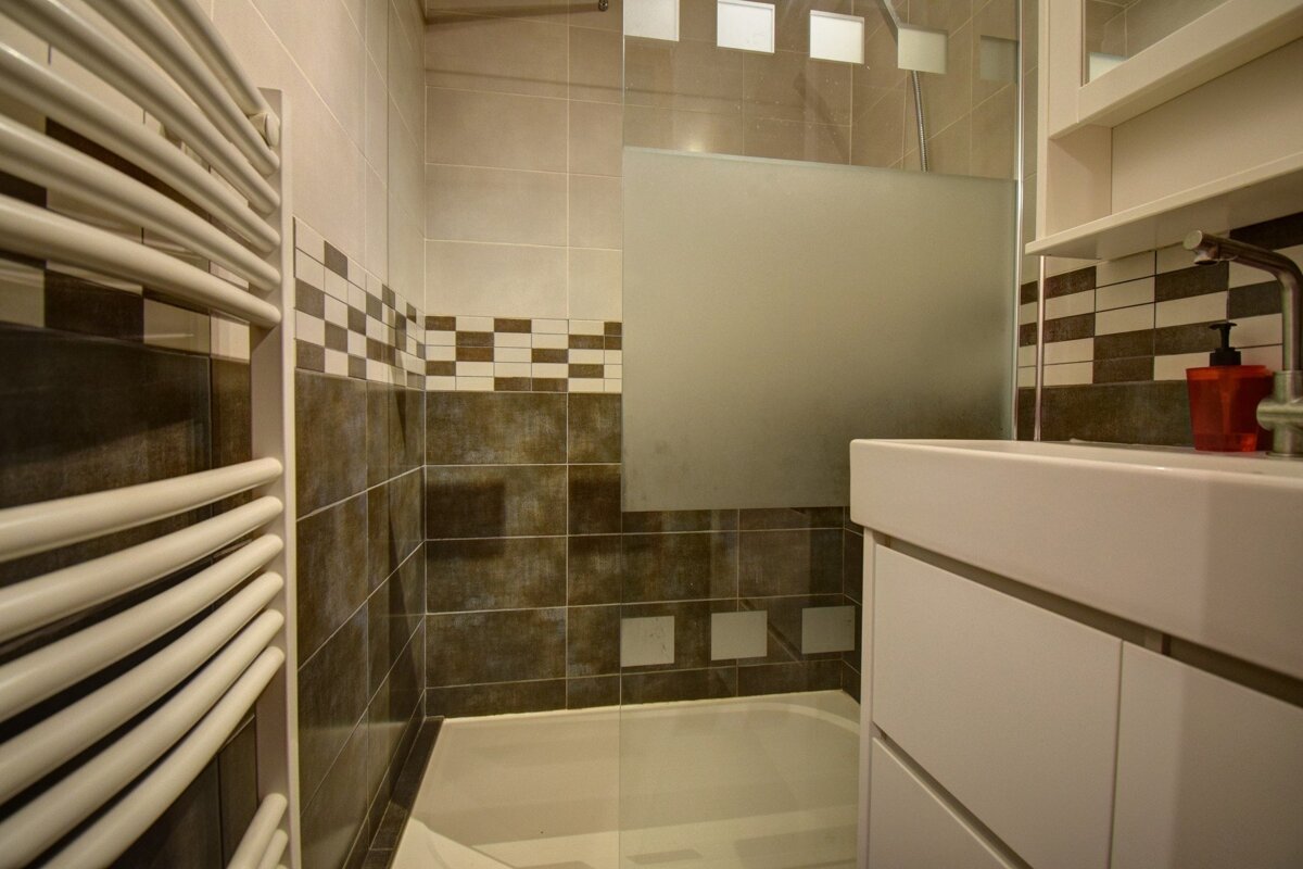 Small modern bathroom with a white towel radiator, vanity sink, and a bathtub with a frosted glass screen.