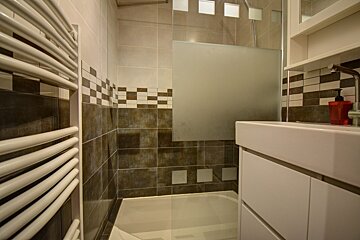 Small modern bathroom with a white towel radiator, vanity sink, and a bathtub with a frosted glass screen.