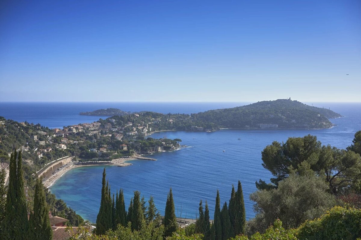 Scenic coastal town along a vibrant blue bay, framed by green hills and cypress trees under a clear sky. A serene Mediterranean vista.