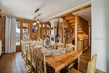 Cozy dining room in a wooden chalet featuring a long set table, rustic chairs, a fireplace, and a wooden staircase.