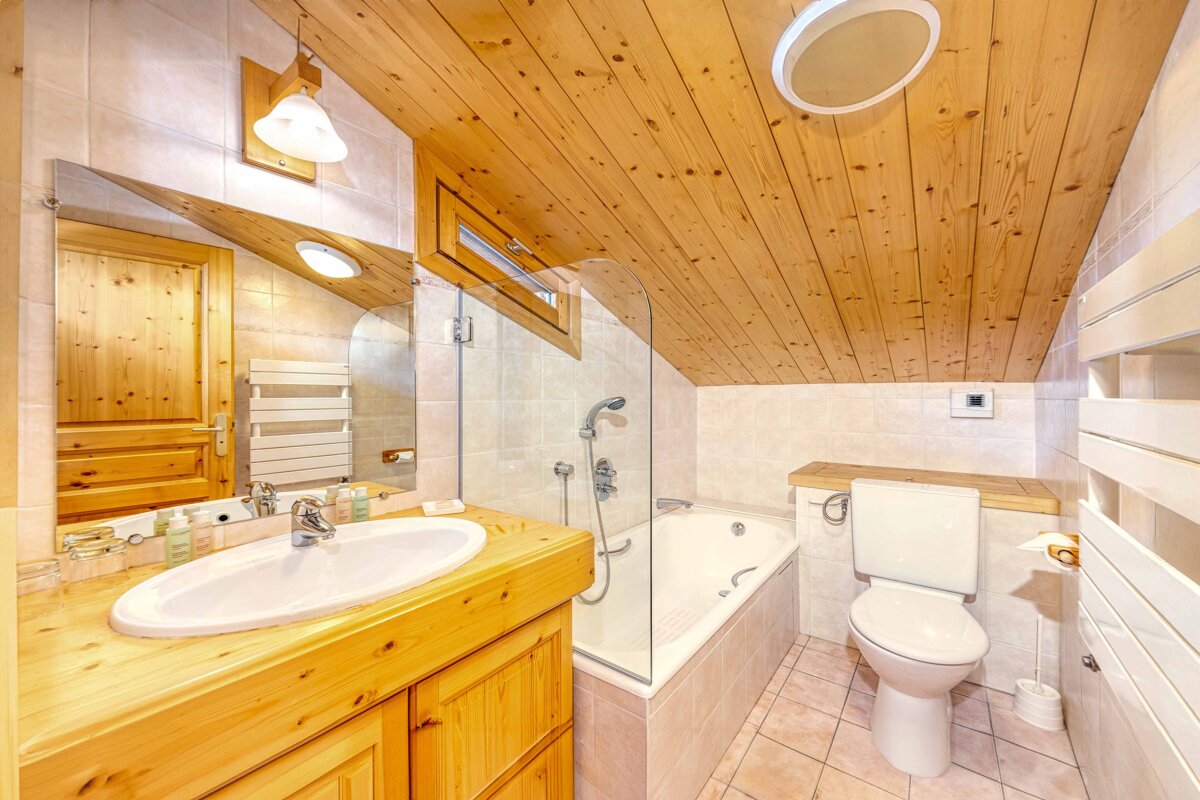 Cozy, cabin-style bathroom featuring slanted wooden ceilings, a matching vanity, a bathtub with a glass partition, and a toilet.