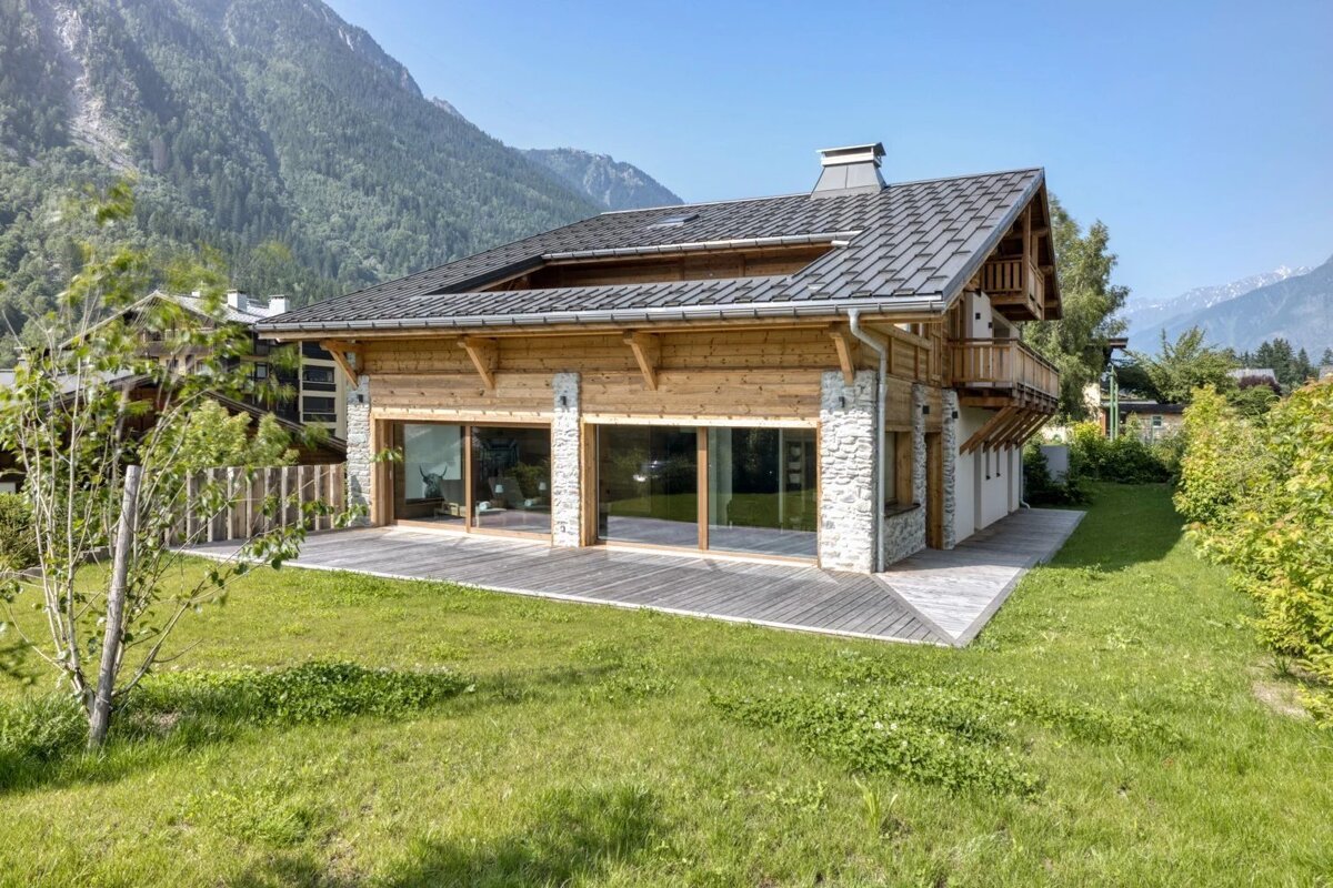 Modern chalet with wood and stone, large windows, a deck, and green lawn, set against a stunning mountain backdrop under a clear sky.