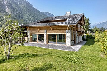 Luxury mountain home with timber and stone facade, expansive windows, a wooden deck, and a lush green yard, framed by majestic peaks.