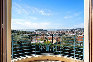 A sunny balcony view of a coastal city with red roofs and green trees, stretching to the blue sea under a clear sky, framed by a blue railing.