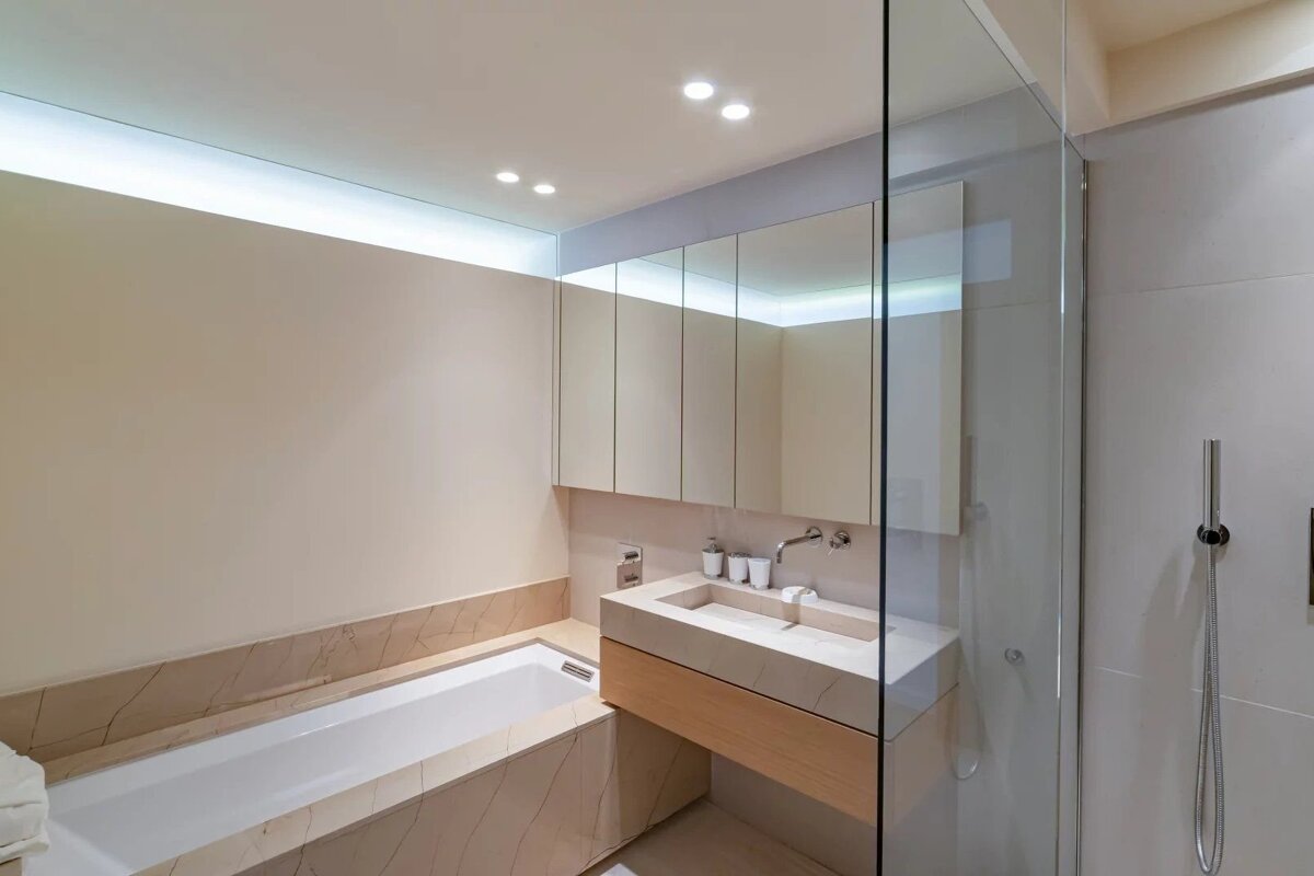 Modern minimalist bathroom in beige tones, featuring a built-in bathtub, a floating wooden vanity, large mirrors, and a glass shower.