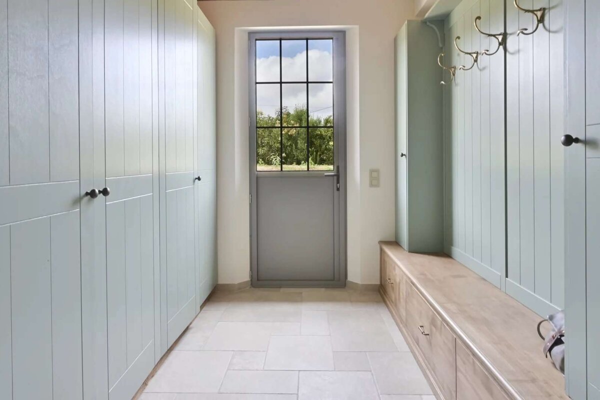 A light green mudroom features built-in full-height cabinets, a wooden storage bench with coat hooks, a tiled floor, and a grey door with gridded glass.
