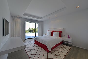 A bright, modern bedroom featuring white walls, a red-accented bed & geometric rug. Large glass doors reveal an outdoor pool and lush landscape.