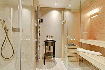A modern bathroom with a walk-in shower, freestanding black sink, and a glass-enclosed sauna featuring light wood interior and heated rocks.