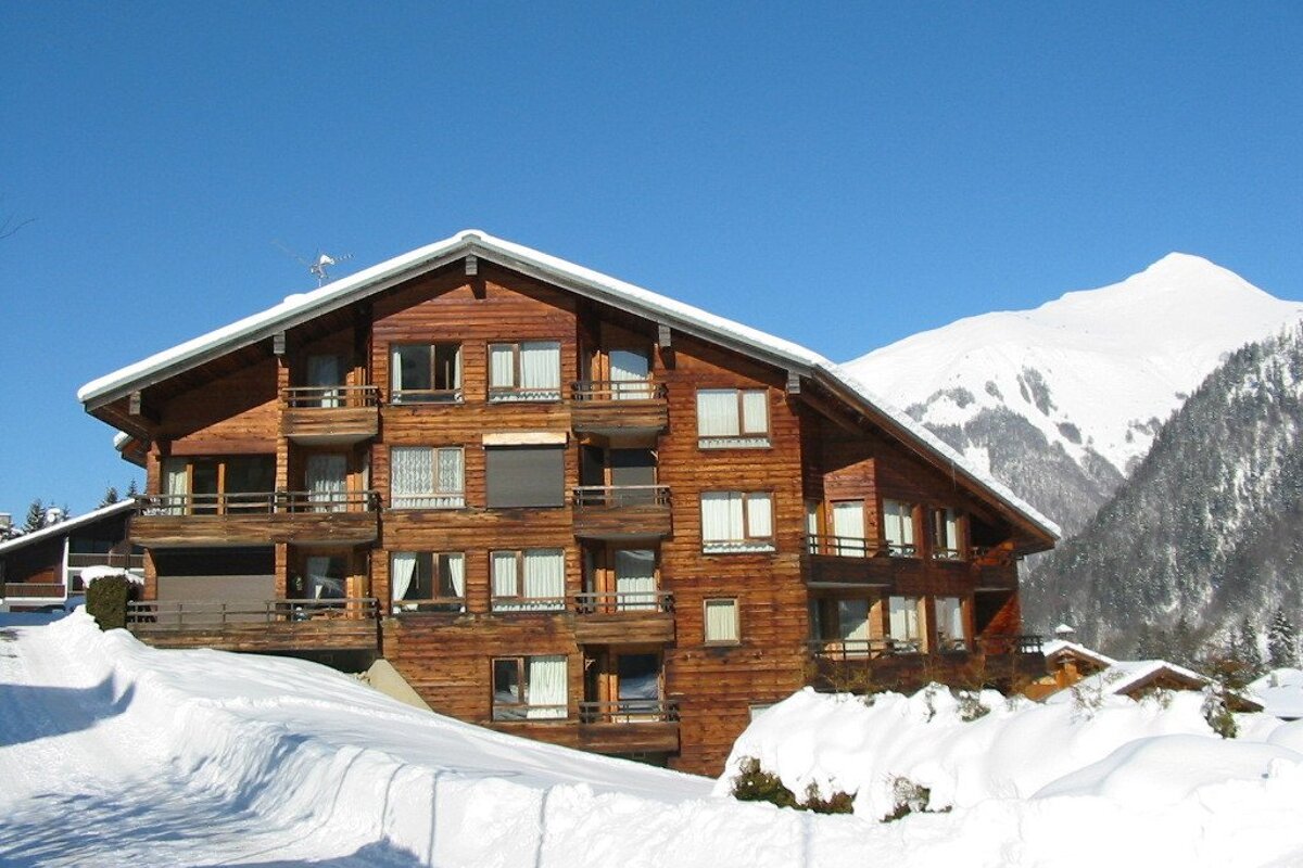 A large multi-story wooden chalet with balconies, surrounded by deep snow, against towering snow-capped mountains under a clear blue sky.