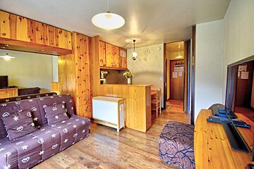 A bright living/dining room with two patterned sofa beds, a wooden dining table and chairs, light blue carpet, and wood wall accents.