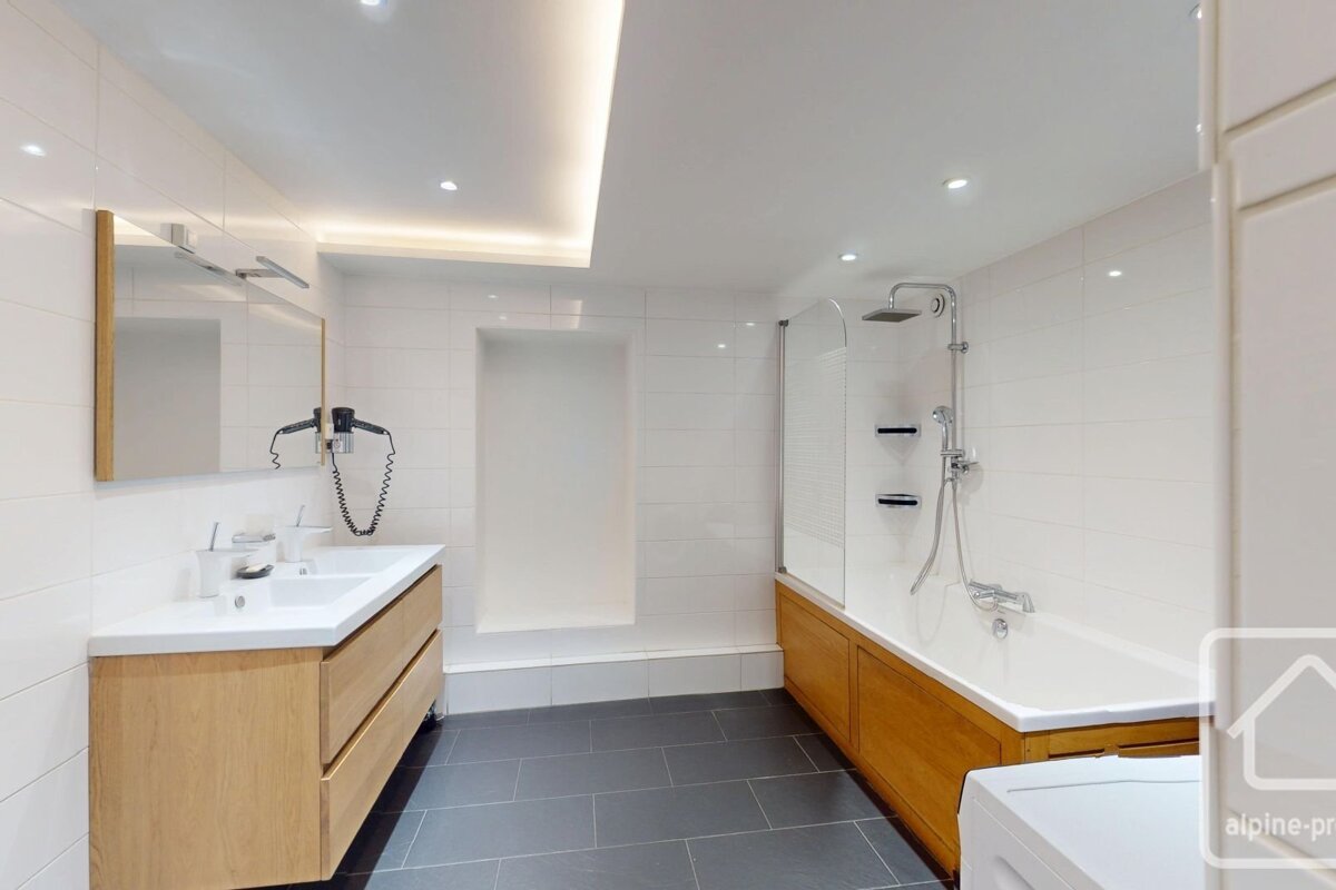 Modern bathroom with white tiled walls, dark gray floor, wooden double vanity, mirror, and a bathtub with a shower and wooden paneling.