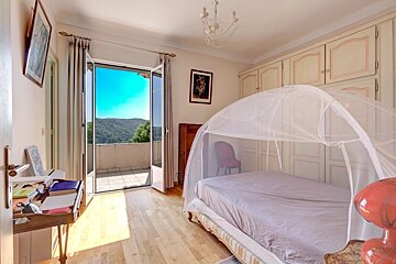Sunny bedroom with a bed covered by a mosquito net, wooden floors, and open balcony doors showing a beautiful scenic view.