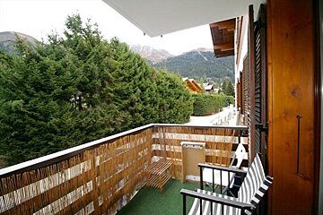 A balcony with a bamboo screen and striped chairs overlooks a dense pine forest and distant mountains.