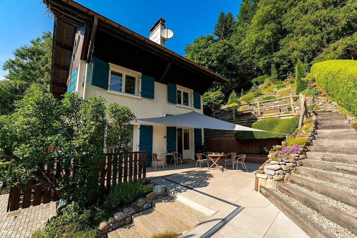 A white house with blue shutters, a shaded patio, and terraced garden steps climbing a lush green hillside under a clear blue sky.