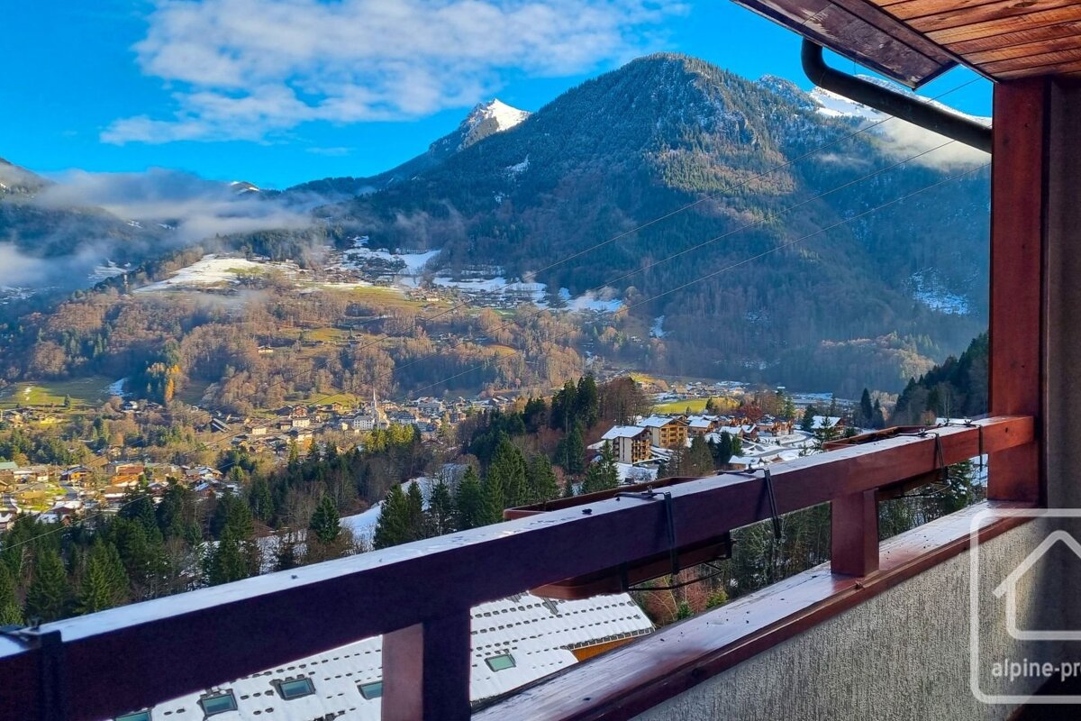 Balcony view overlooking a snowy mountain village and valley, with towering peaks under a blue sky.