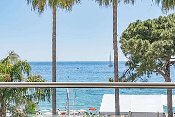 Sunny coastal view from a balcony with palm trees framing the blue sea, boats, and a beach with umbrellas and white tents.