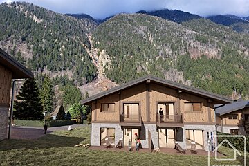A digital rendering shows alpine-style chalets with stone and wood facades nestled against a steep, pine-forested mountain.