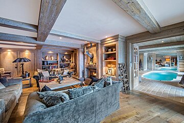 A cozy, rustic wooden living room with a fireplace, deer head, and plush couches, opening onto an indoor pool area with two pools.