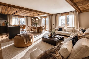 Cozy alpine chalet interior featuring wooden ceilings, plush sofas with fur throws, a leather ottoman, and a sunlit dining area.