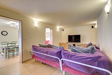 A cozy living room features a purple sofa facing a TV, light wood floors, and wall lights. A dining area with green chairs is visible through an open doorway.