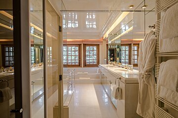 A pristine white bathroom features a double vanity, large windows, a spacious tub, and a plush robe. Mirrors enhance the bright, luxurious ambiance.