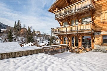A wooden chalet with balconies and a snow-covered terrace overlooking a winter mountain landscape.