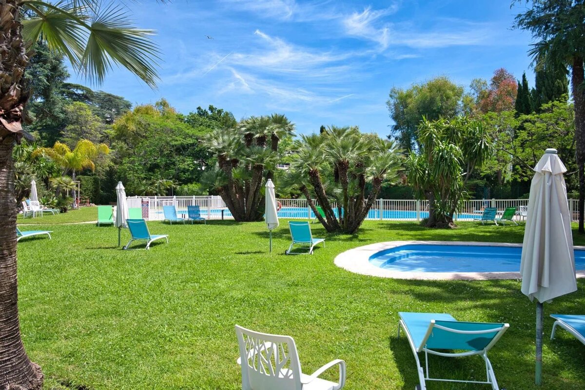 A sunny resort garden features two blue swimming pools, a lush green lawn, various lounge chairs, white umbrellas, and palm trees under a clear sky.