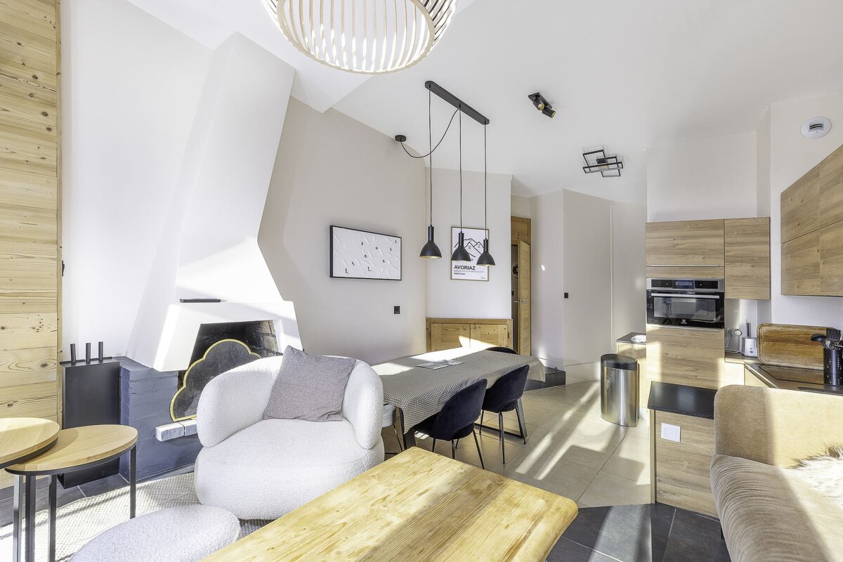Modern chalet interior with a white fireplace, round armchair, wooden dining table, and light wood kitchen cabinets.