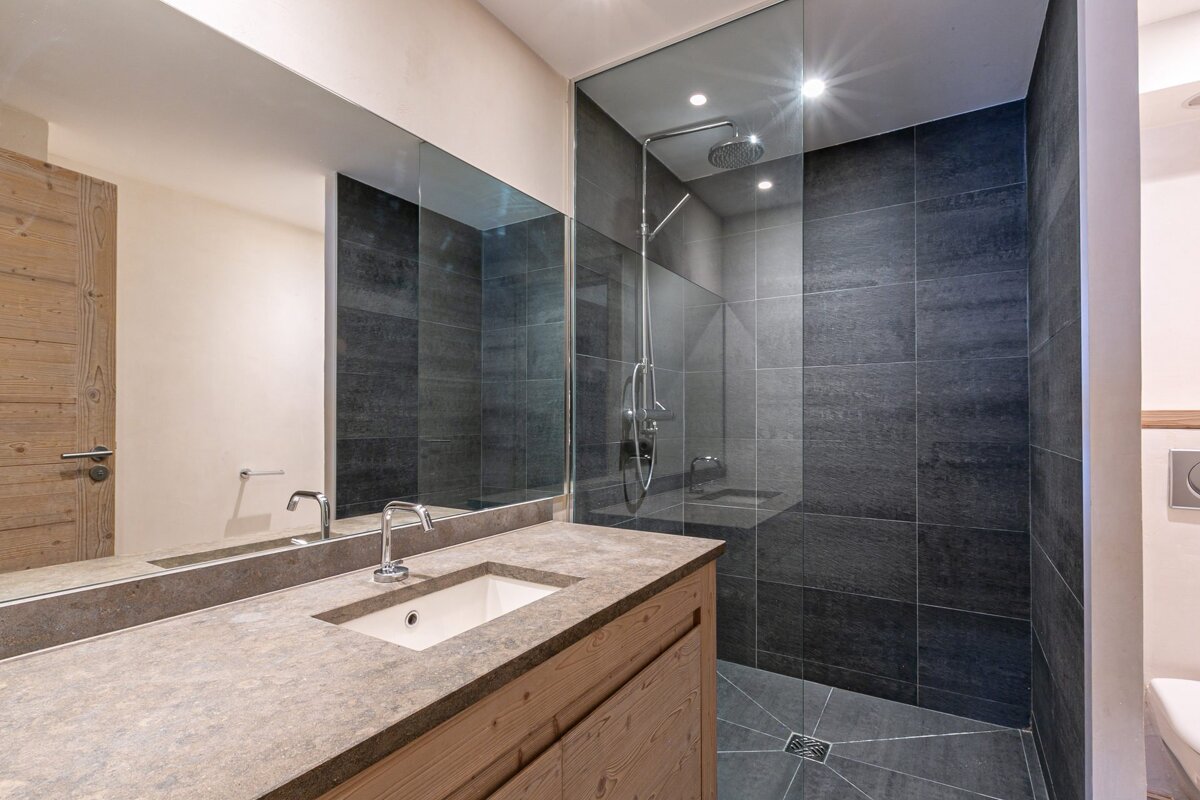 Modern bathroom with a dark tiled walk-in shower, glass partition, stone vanity, integrated sink, and light wood cabinet.
