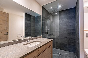 Modern bathroom with a dark tiled walk-in shower, glass partition, stone vanity, integrated sink, and light wood cabinet.
