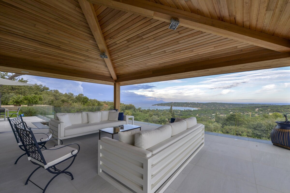 Spacious outdoor patio with modern white furniture under a wooden ceiling, boasting a panoramic view of a bay, sea, and lush green landscape.