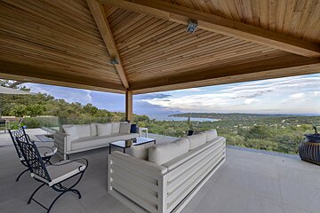 Spacious outdoor patio with modern white furniture under a wooden ceiling, boasting a panoramic view of a bay, sea, and lush green landscape.