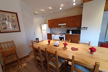 A cozy dining area featuring a long wooden table and chairs, with a rustic kitchen in the background.