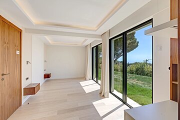 An empty, modern room with light wood floors and large windows looking out onto a bright green garden.