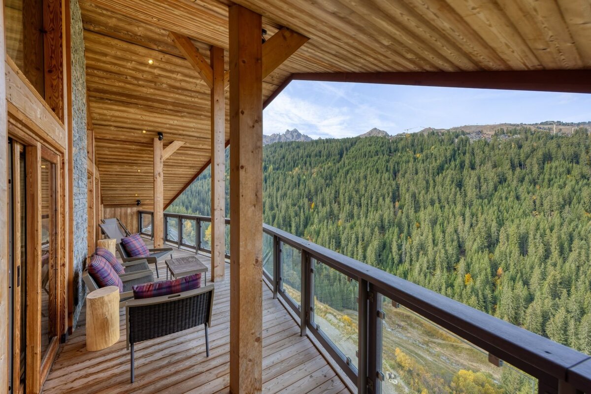 A rustic wooden balcony with outdoor seating overlooks a vast pine forest and majestic mountains under a blue sky, offering stunning views.