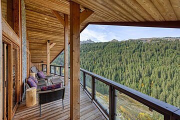 A rustic wooden balcony with outdoor seating overlooks a vast pine forest and majestic mountains under a blue sky, offering stunning views.