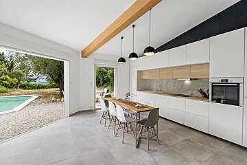 Modern white kitchen with a wooden dining table, black pendant lights, and large sliding doors opening to an outdoor swimming pool.