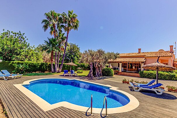 A sunny villa with a blue swimming pool, wooden deck, sun loungers, palm trees, and thatched umbrellas, surrounded by lush greenery.