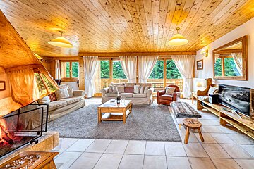 A cozy, wood-paneled living room features a crackling fireplace, plush sofas, and large windows overlooking a vibrant forest.