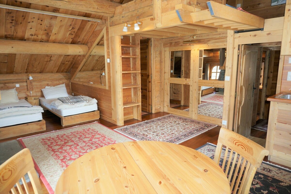 A cozy wooden cabin interior featuring twin beds, timber beams, patterned rugs, and a dining table with chairs.
