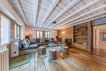 A warm, rustic living room with an L-shaped couch, beamed wood ceiling, and hardwood floor. Features built-in shelving and natural wood accents.