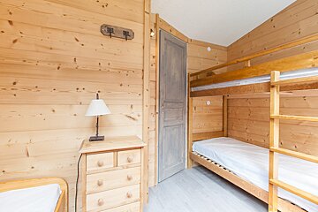 A cozy, rustic bedroom featuring light wood paneled walls, a wooden bunk bed, a dresser with a lamp, and a gray door.