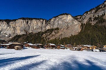 A snow-covered village with wooden chalets sits at the base of towering rocky cliffs and a dense pine forest under a bright blue sky.