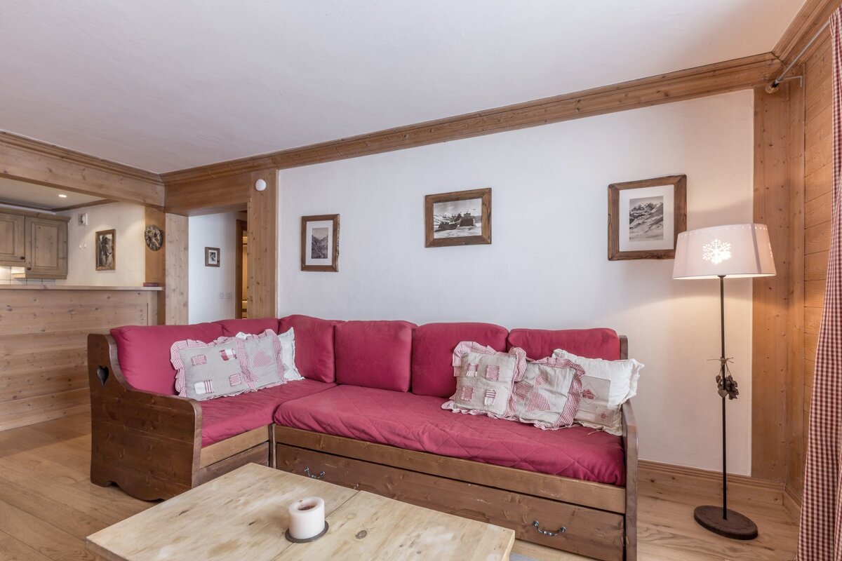 A cozy, chalet-style living room features a red L-shaped couch, wooden paneling, white walls, and framed mountain art. A snowflake lamp adds charm.